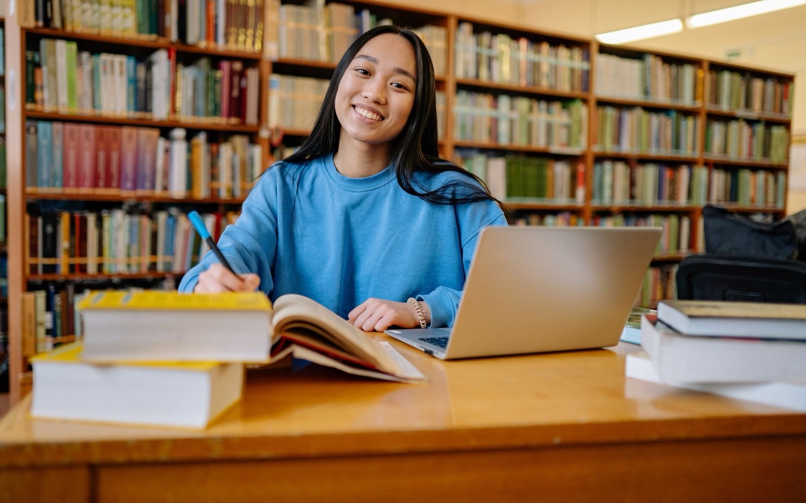 A sixth-form student with books and a laptop at a library desk