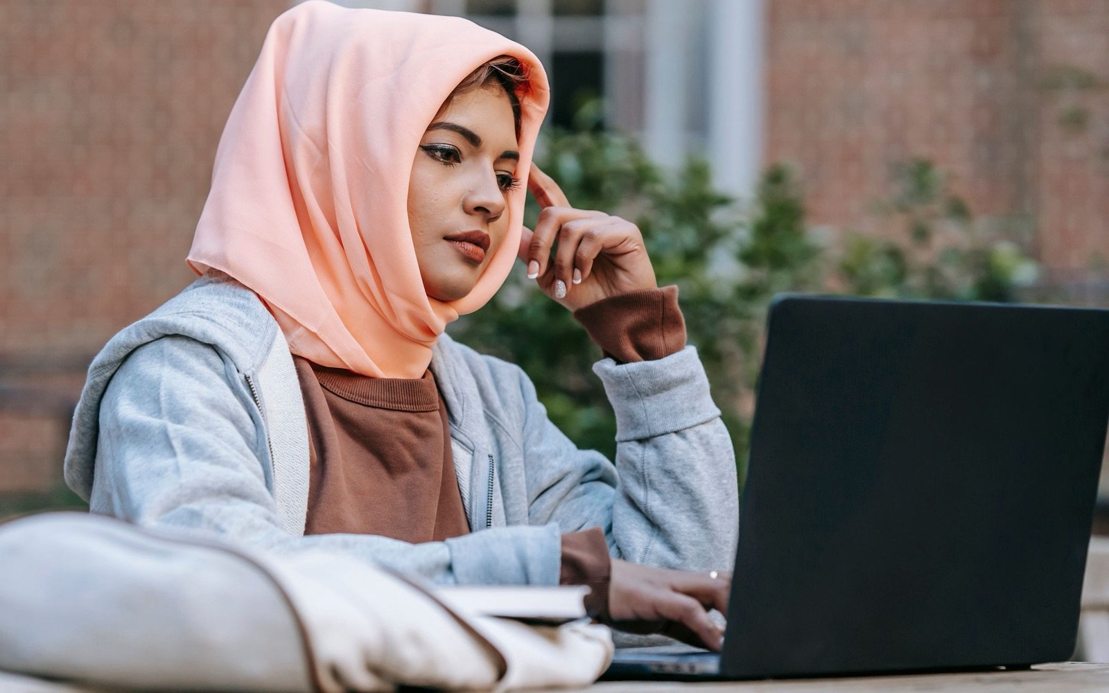 A student in a headscarf thinking while she works on her laptop outdoors