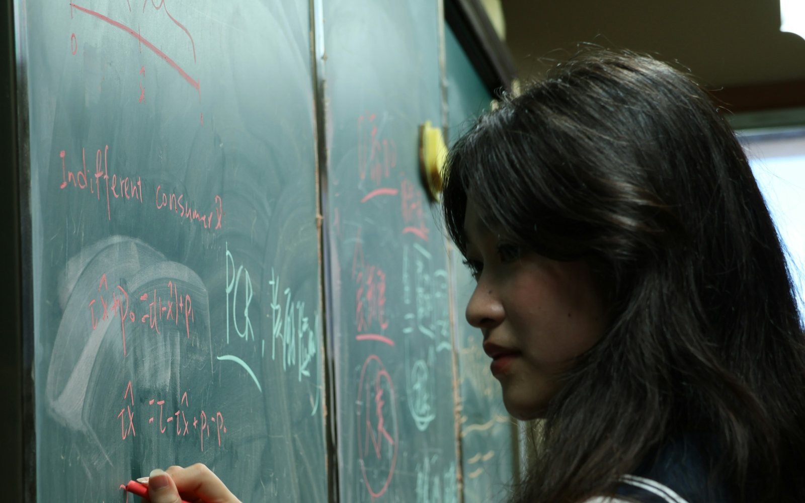 A sixth-form student in school uniform working at a chalkboard covered in equations