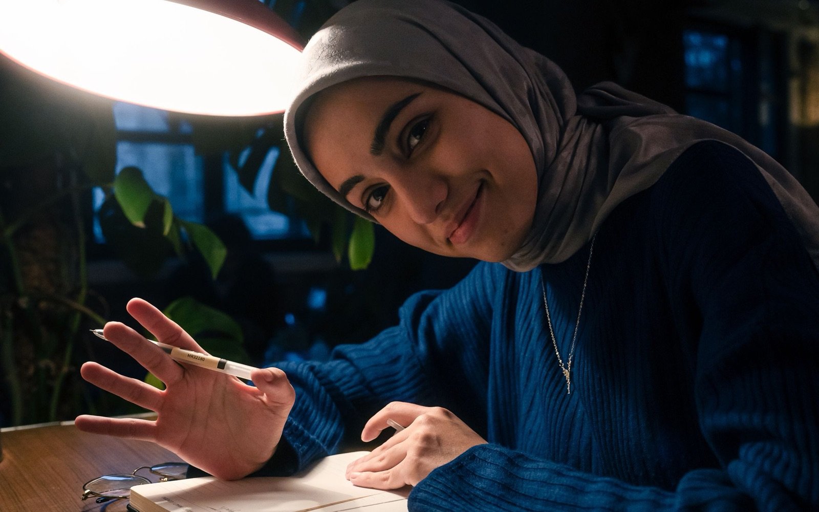 A student in a headscarf smiling as she studies at a desk at home in the evening