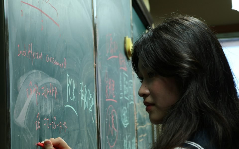 A sixth-form student in school uniform working at a chalkboard covered in equations