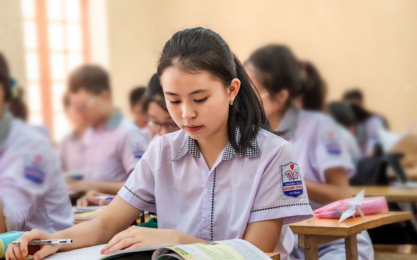 A student in school uniform concentrating on her work in a busy classroom